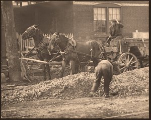 Horizontal, black and white photograph of a horse-drawn wagon at a construction site. A man shovels dirt and rock while another drives the wagon